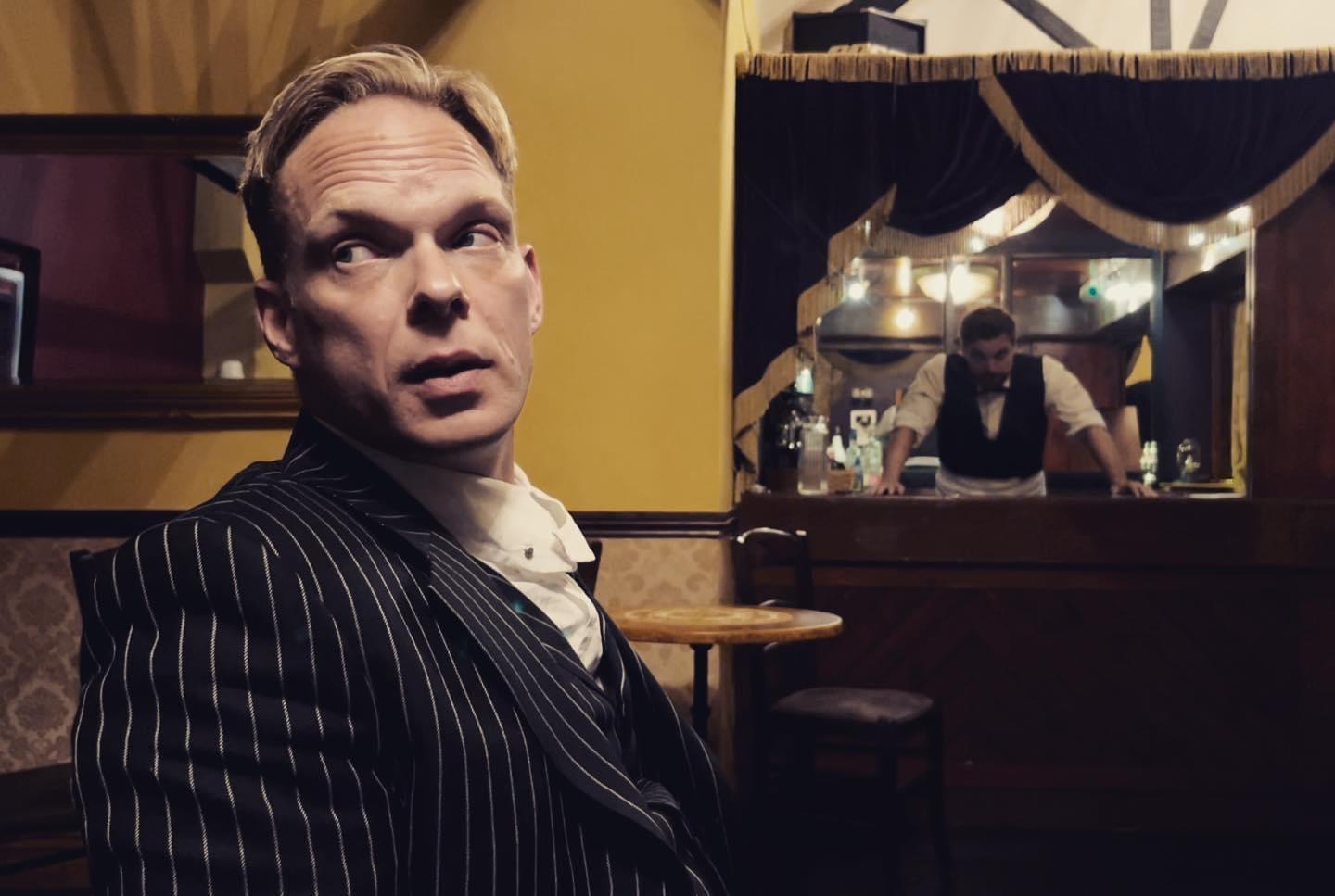 Christian Steel in character on screen, pinstripe suit in a warm-lit bar